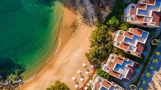Vista aérea de un hotel frente al mar con varias construcciones, una playa de arena y aguas azules cristalinas.