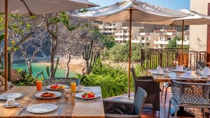 Un área de comedor al aire libre con mesas y sillas, vista a la playa y cactus.