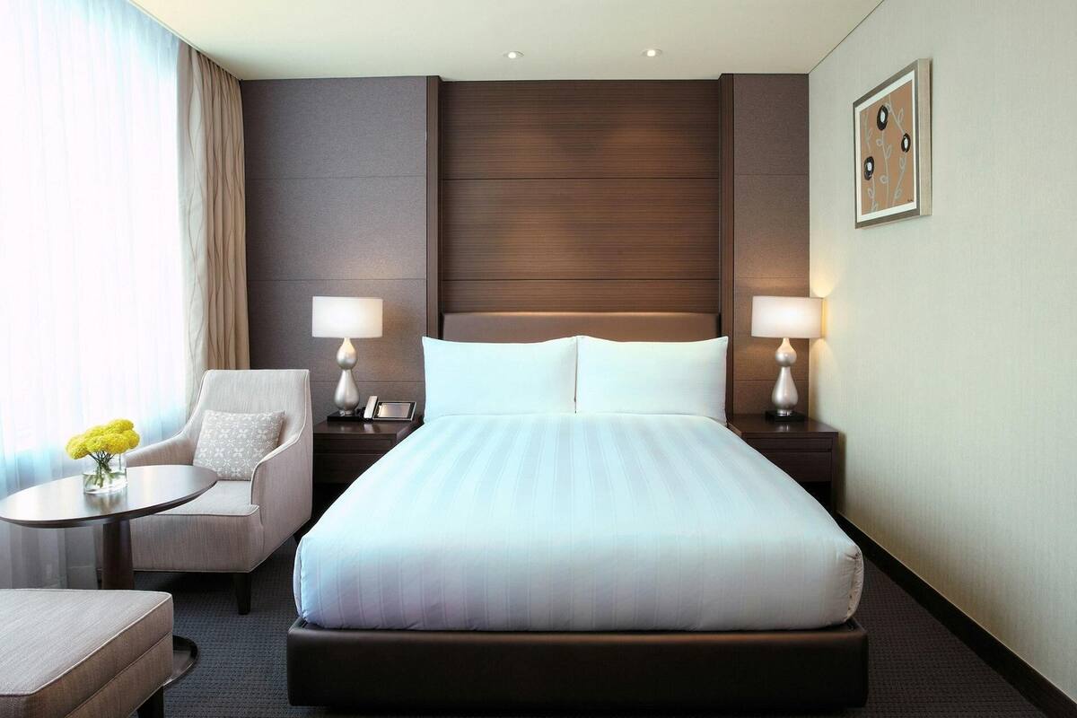 Une chambre d’hôtel avec un grand lit, deux tables de chevet avec des lampes, une chaise, une petite table sur laquelle est posé un vase de fleurs et un tableau accroché au mur.