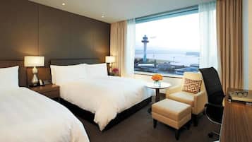 A hotel room with two beds, a small table with a vase, a chair, and a view of a tower through a window.