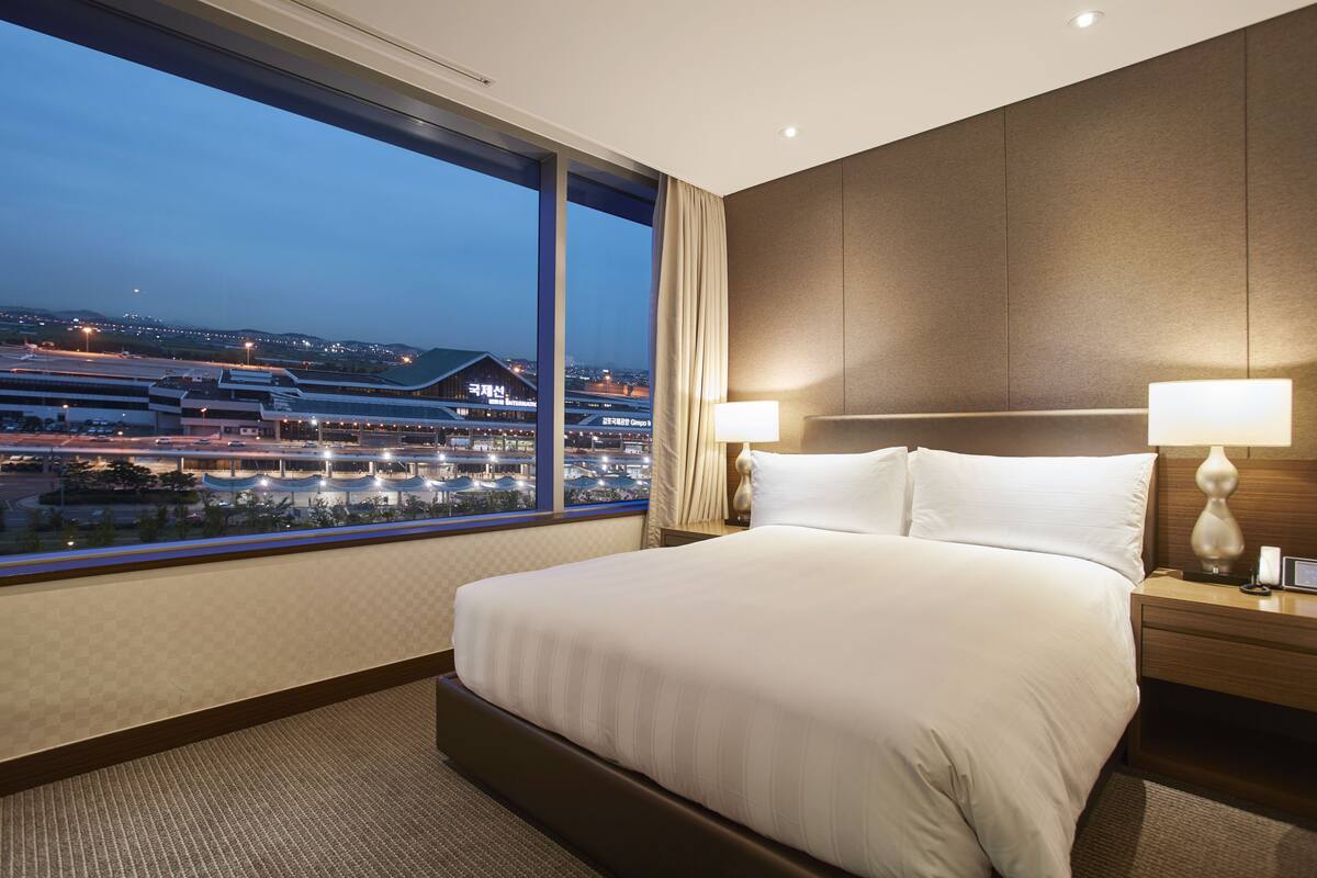 Une chambre d’hôtel dotée d’un grand lit, de deux tables de chevet, d’une commode, d ’une fenêtre donnant sur un aéroport et sur le paysage urbain, et présentant un aménagement intérieur moderne.