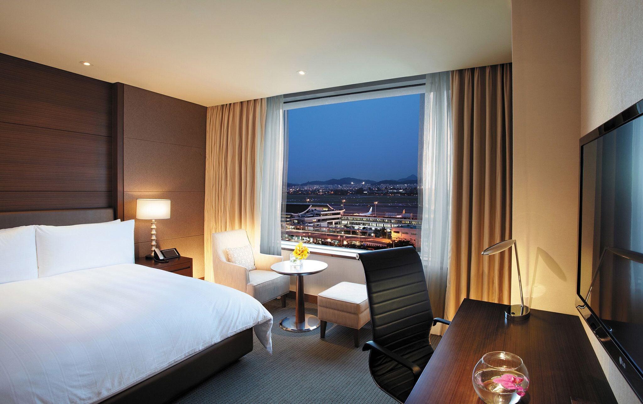 A hotel room with a bed, a desk, a chair, a small table, and a TV.