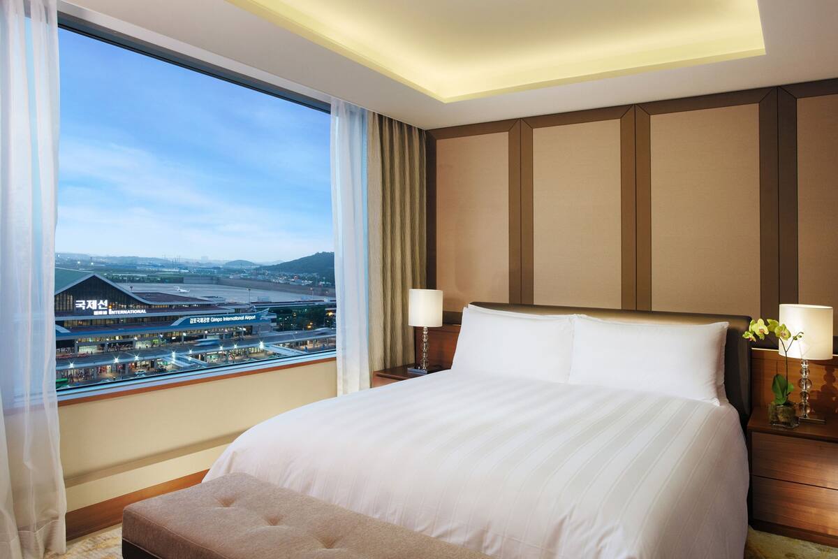 Une chambre d’hôtel avec un grand lit, une vue sur un aéroport et une table de chevet avec une lampe.