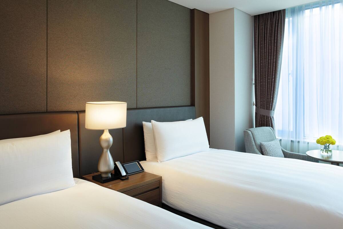 Une chambre d’hôtel avec deux lits, une table de chevet avec une lampe, une chaise, une petite table avec des fleurs et une vue sur l’extérieur à travers des rideaux légers.