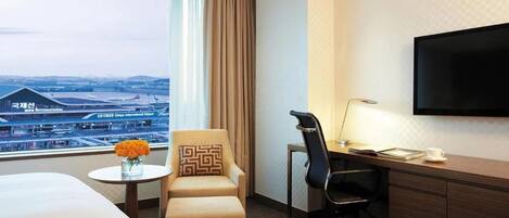 A hotel room with a bed, a desk, a chair, a TV, a small table with a vase of flowers, and a view of an airport.