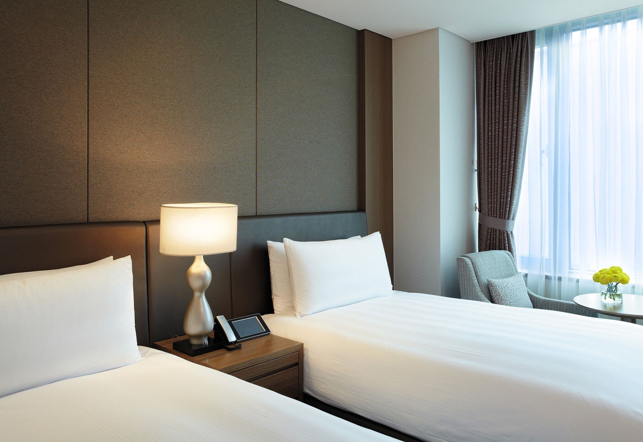 A hotel room with two beds, a bedside table with a lamp, a phone, and a small table with a vase of flowers.