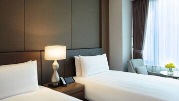 A hotel room with two beds, a bedside table with a lamp, a phone, and a small table with a vase of flowers.