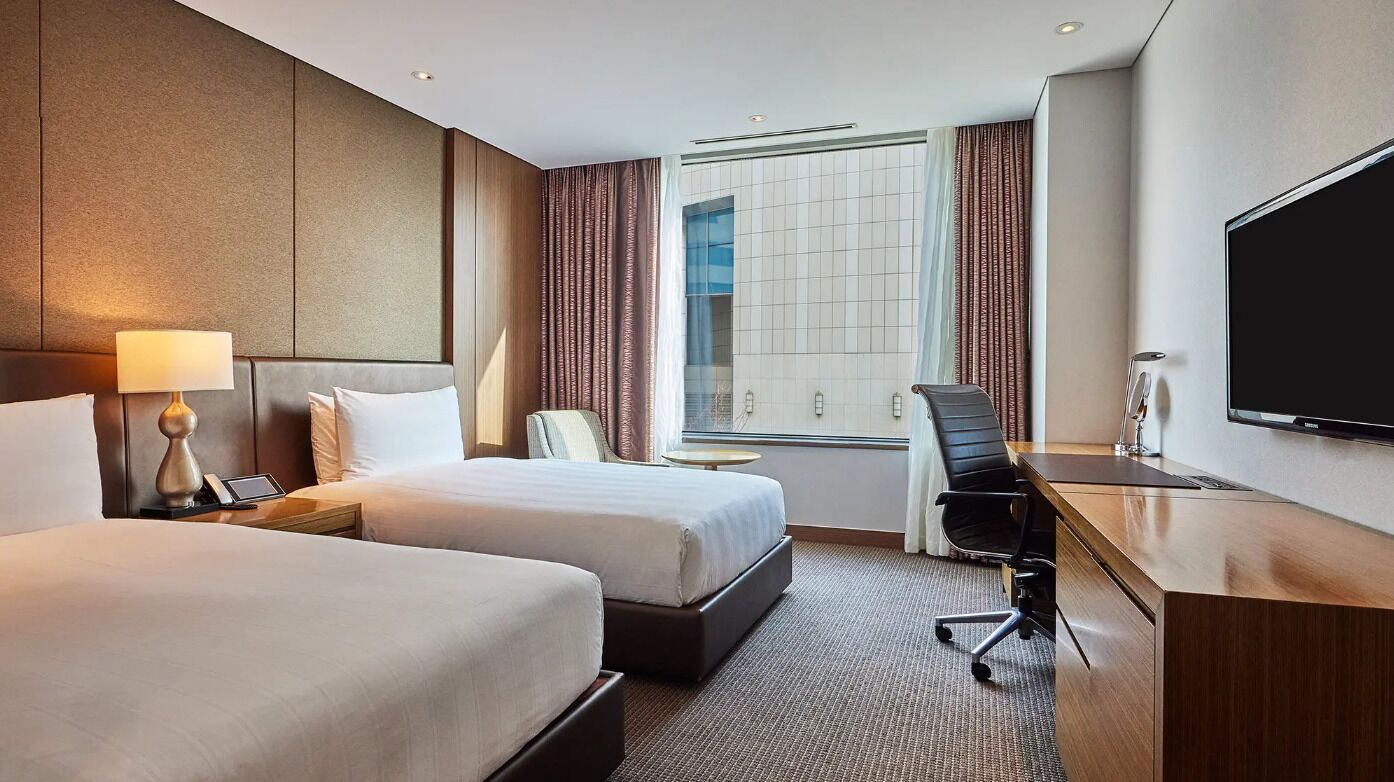 Ein Hotelzimmer mit zwei Betten, einem Schreibtisch, einem Stuhl, einem Fernseher und einem Fenster mit Vorhängen.