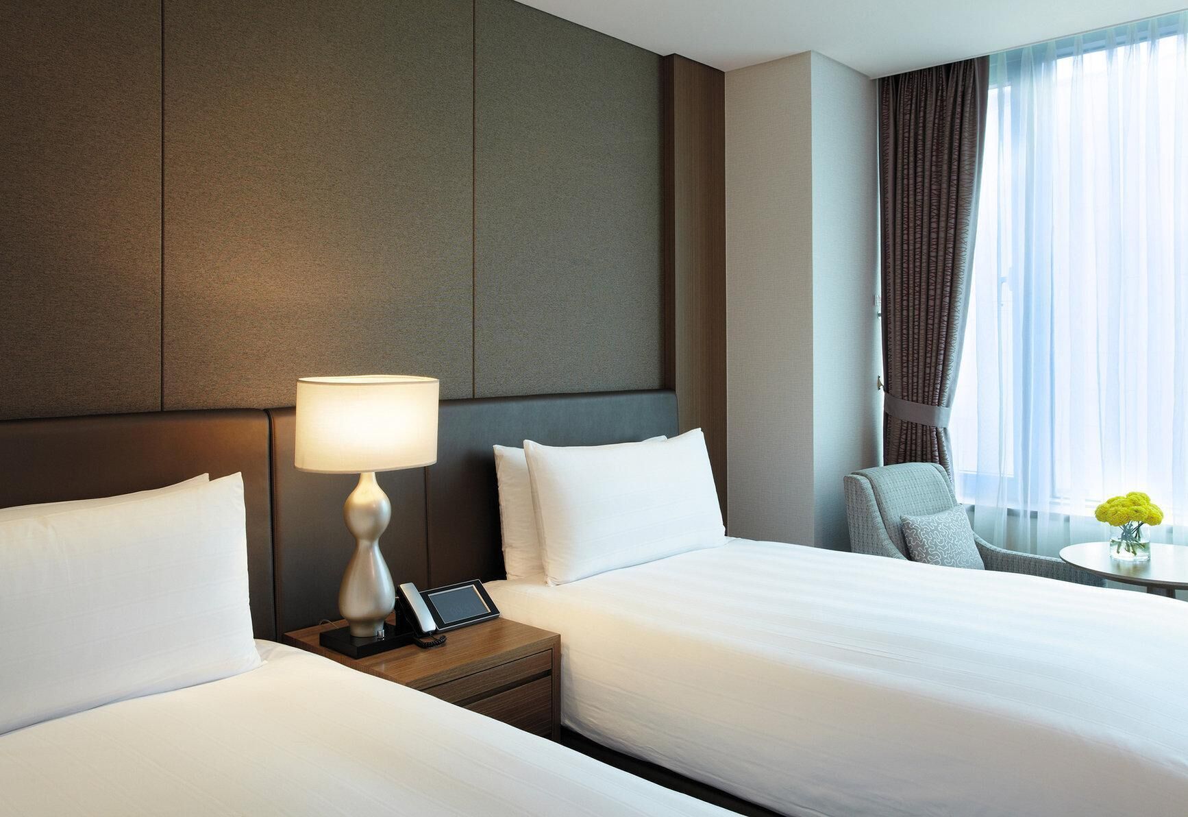 A hotel room with two beds, a bedside table with a lamp, a chair, a small table with a vase, and a view of the outdoors.