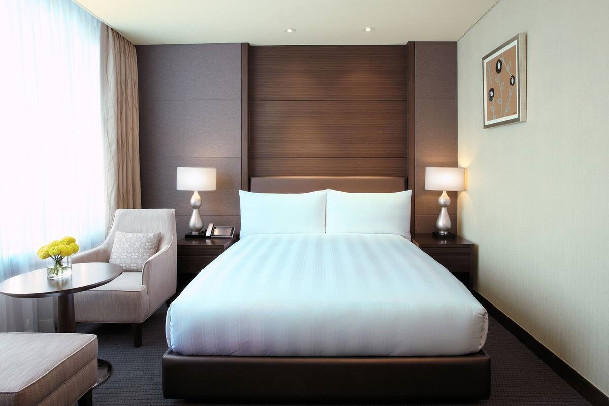 Une chambre d’hôtel avec un grand lit, deux tables de chevet avec des lampes, une chaise et une petite table sur laquelle est posé un vase de fleurs.