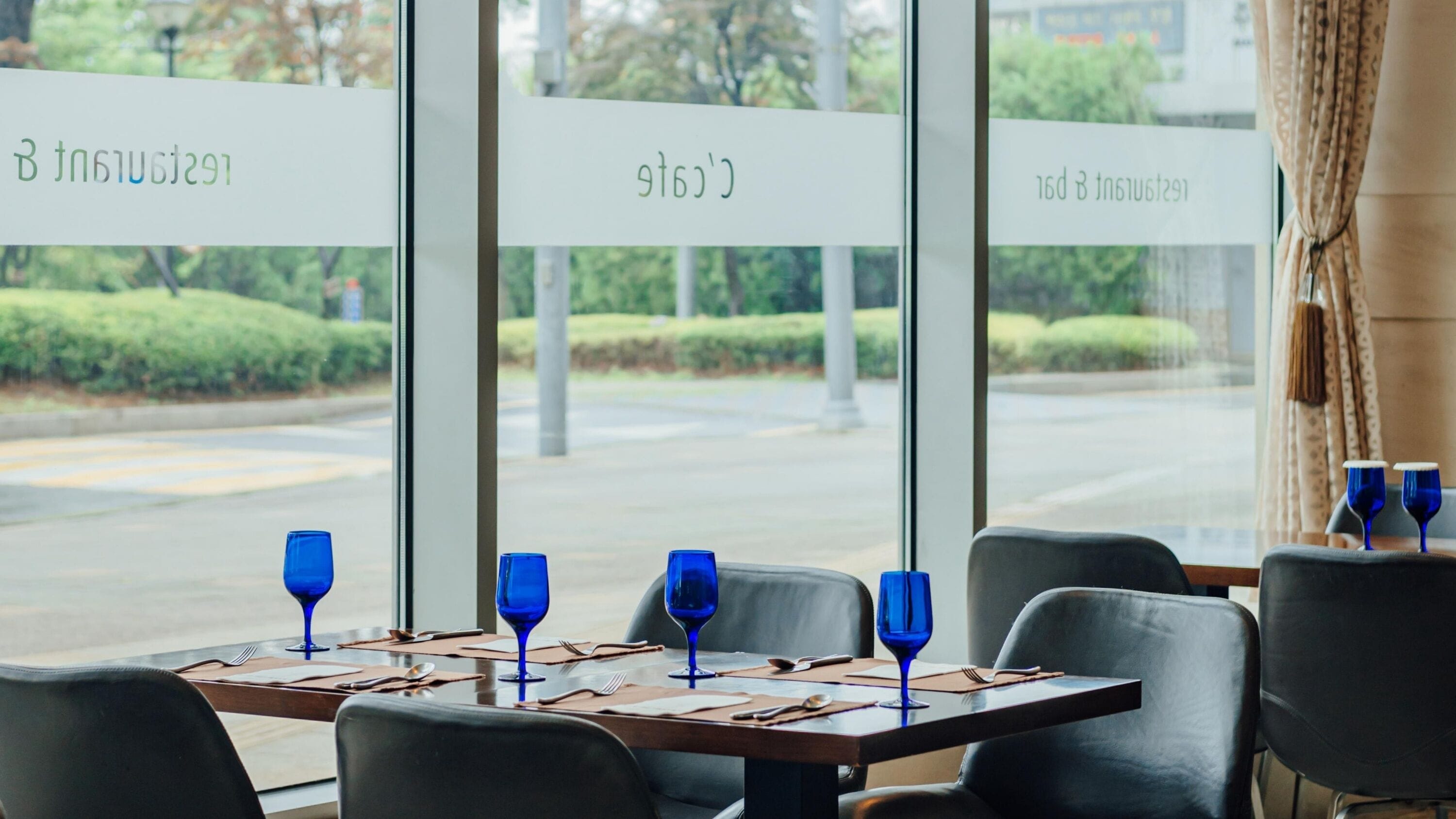 Un ristorante con tavoli apparecchiati, bicchieri di vino blu e vista su una strada e del verde circostante.