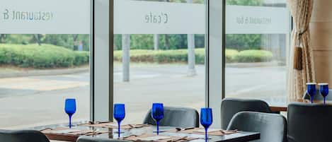 Un ristorante con tavoli apparecchiati, bicchieri di vino blu e vista su una strada e del verde circostante.