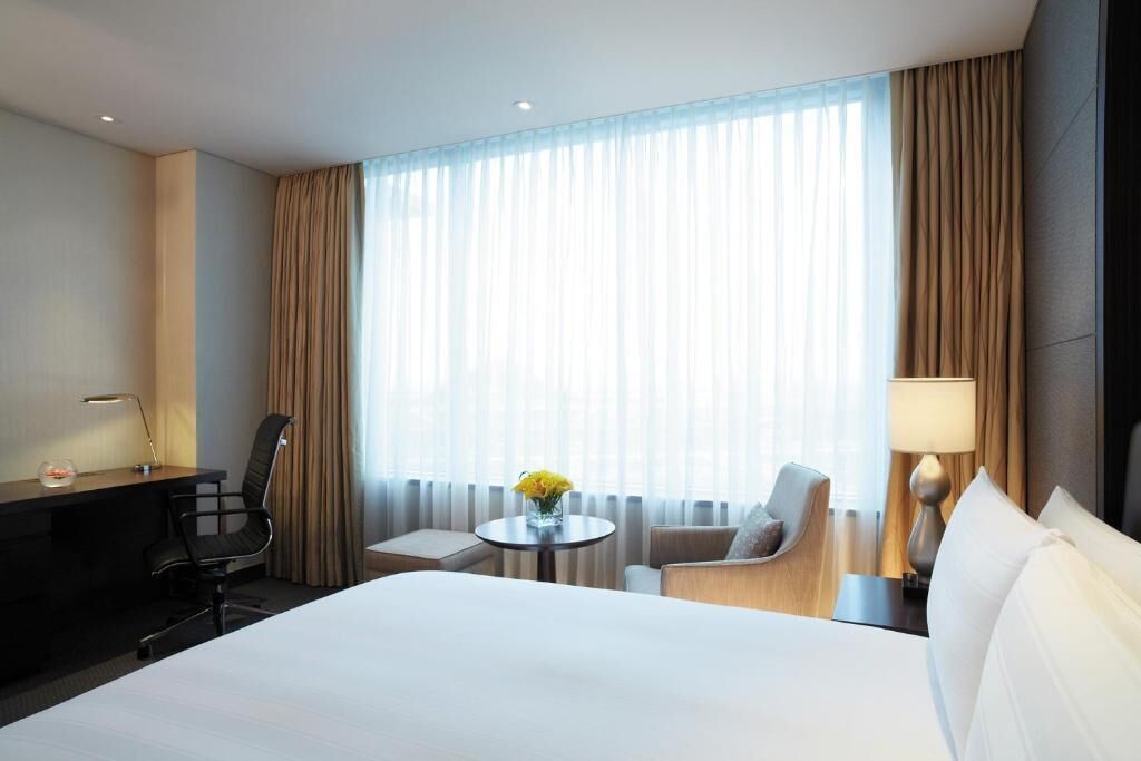 A hotel room with a bed, a desk, a chair, a small table with a vase, and a lamp.