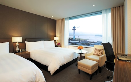 A hotel room with two beds, a small table with a vase of flowers, a chair, and a view of a cityscape and water.