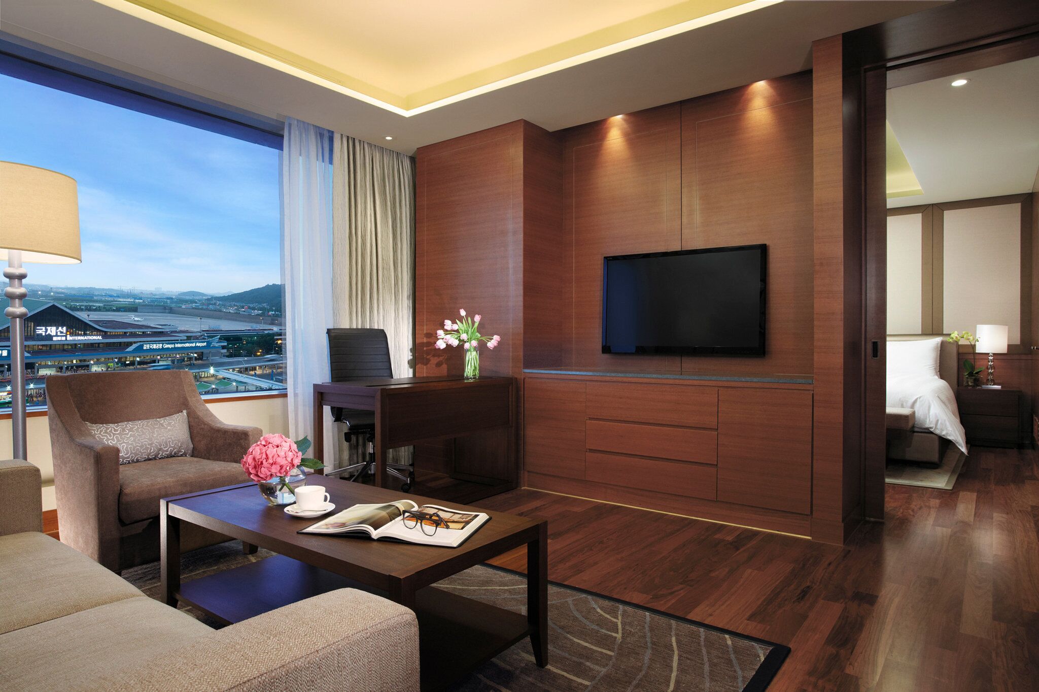 Una habitación de hotel moderna con un amplio ventanal que ofrece vistas a la ciudad, un televisor de pantalla plana montado en la pared, una zona de estar cómoda con una mesita de café y una pequeña mesita auxiliar con un jarrón de flores.