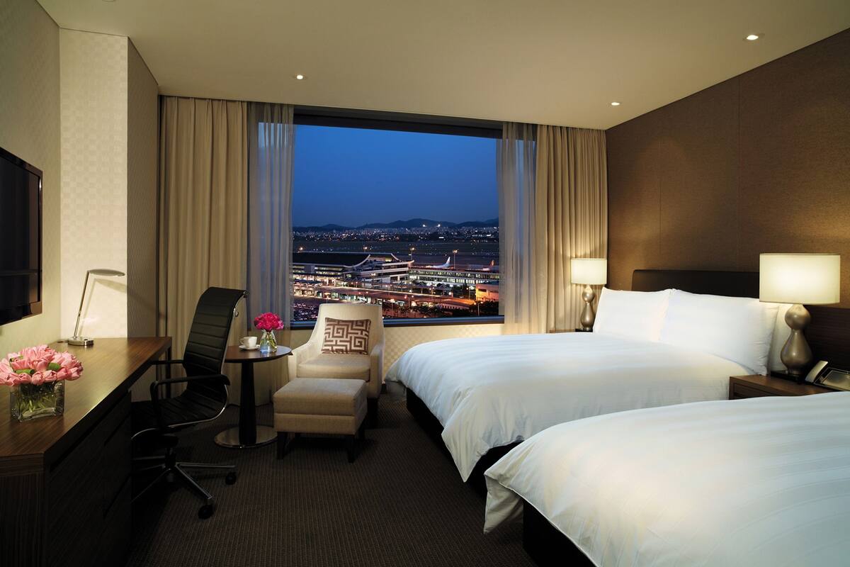 Une chambre d’hôtel avec deux lits, un bureau, une chaise et une vue sur le paysage urbain au crépuscule.