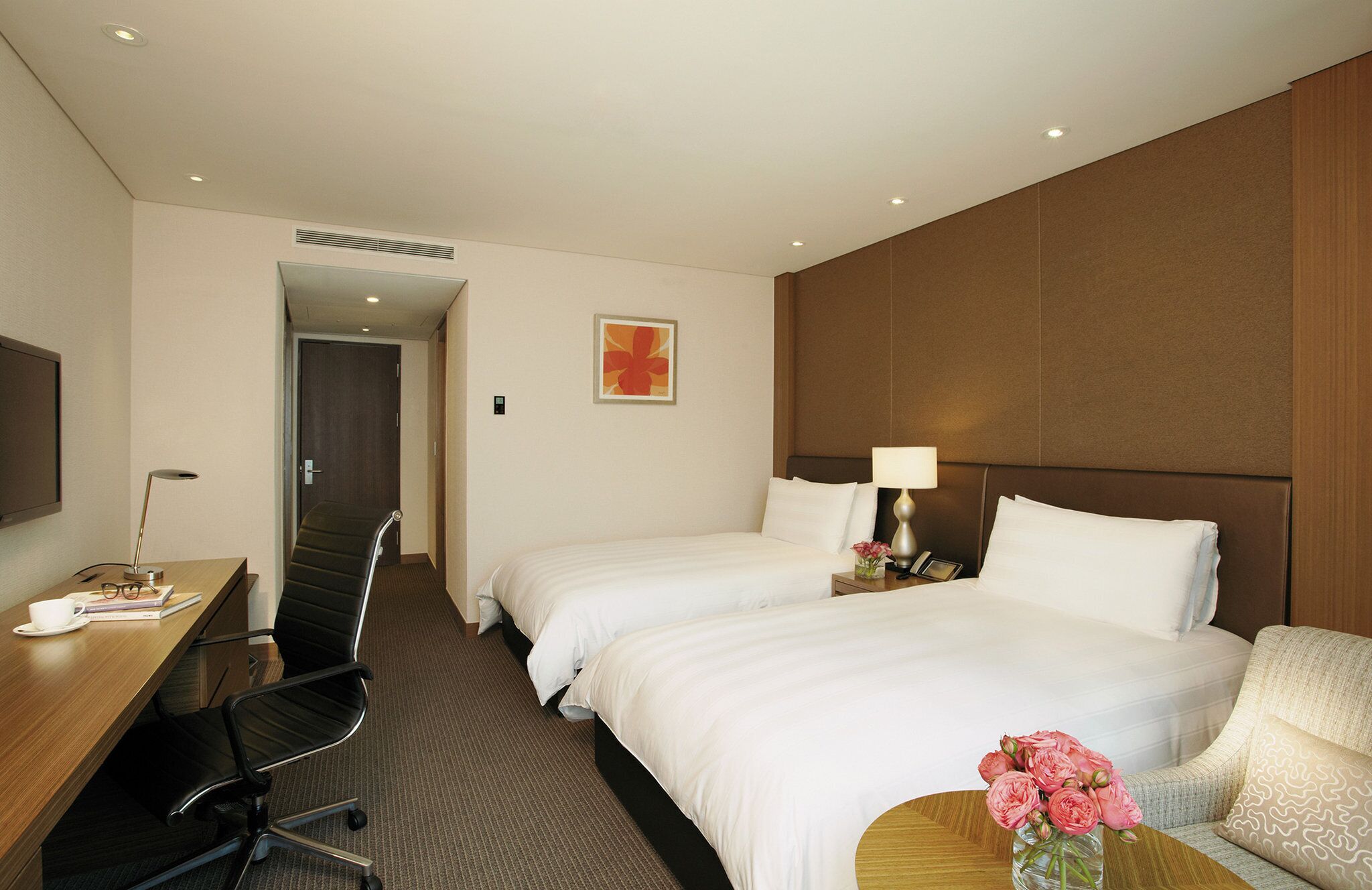 Habitación de hotel con dos camas, un escritorio, una silla, una lámpara y un florero con flores.