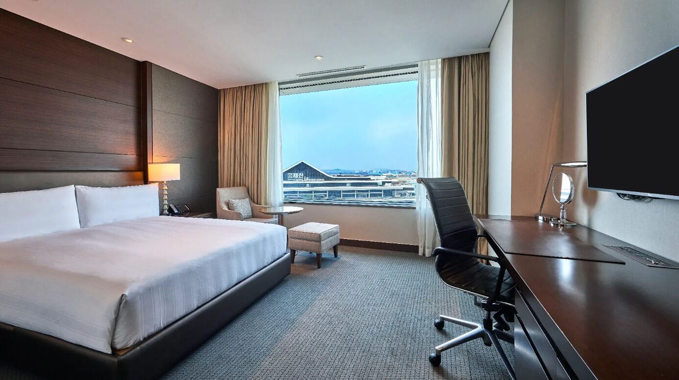 Ein Hotelzimmer mit einem großen Bett, einem Schreibtisch, einem Sessel, einem Fernseher und Blick auf die Stadt.