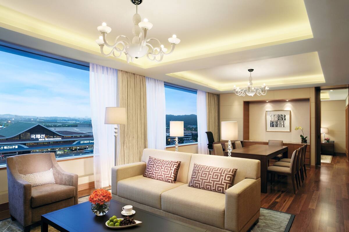 Une chambre d’hôtel équipée d’un canapé, d’un fauteuil, d’une table à manger et de chaises. Une grande fenêtre offre une vue sur un aéroport.
