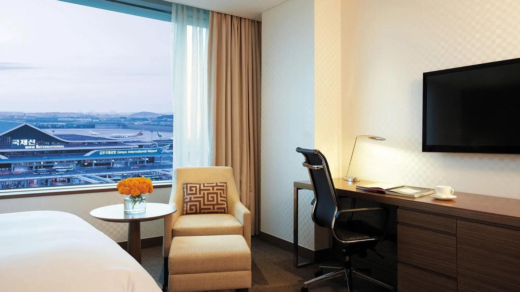 A hotel room with a bed, a desk, a chair, a TV, a window with a view of an airport, and a vase with flowers.