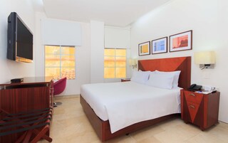 A hotel room with a bed, bedside table, desk, chair, TV, and artwork on the wall.