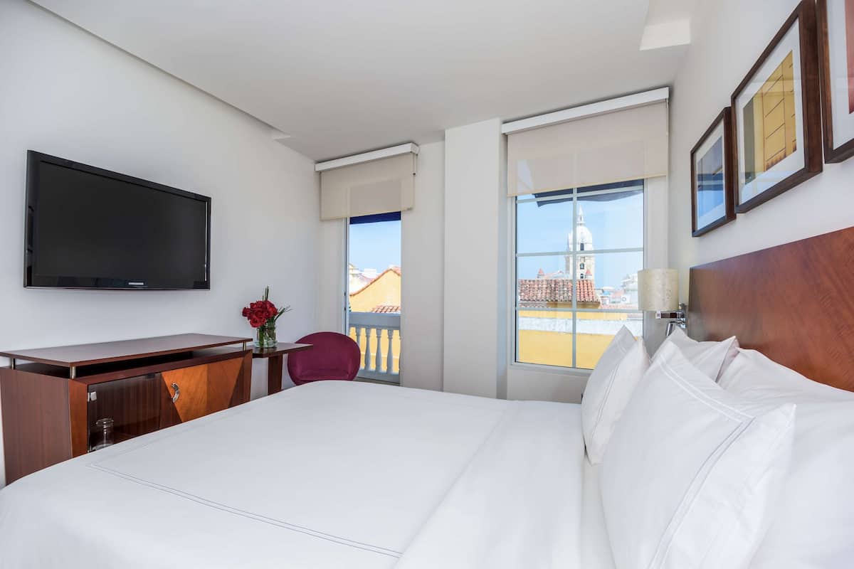 A hotel room with a bed, a desk, a TV, a window with a view of a building, and a chair.