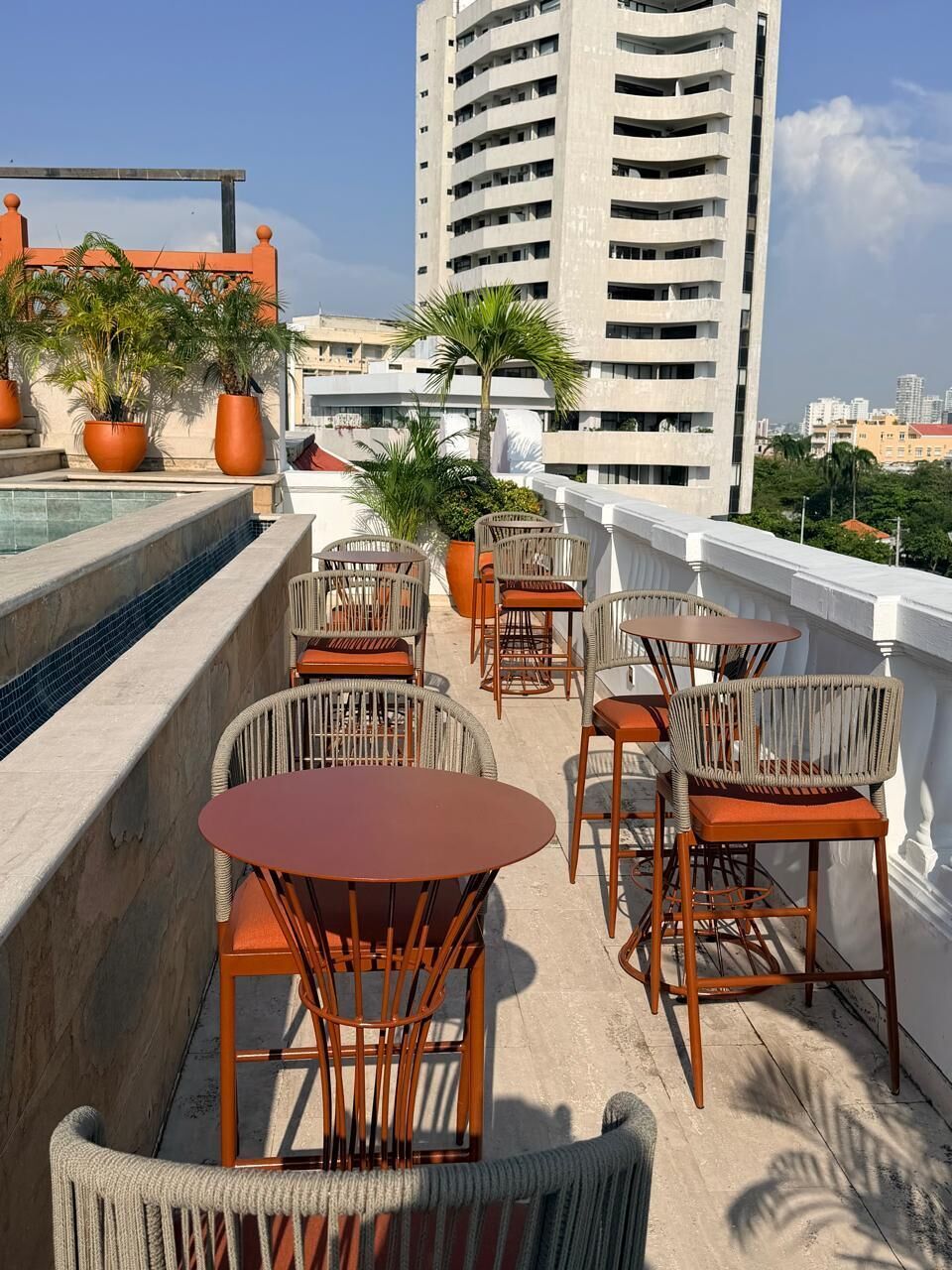 Una terraza en la azotea con mobiliario exterior, una piscina y un edificio alto al fondo.