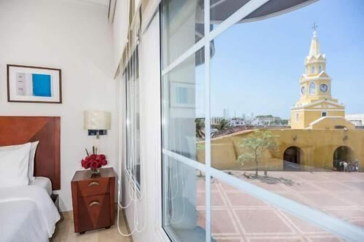 A hotel room with a bed, bedside table, lamp, and a window with a view of a yellow building with a clock tower.