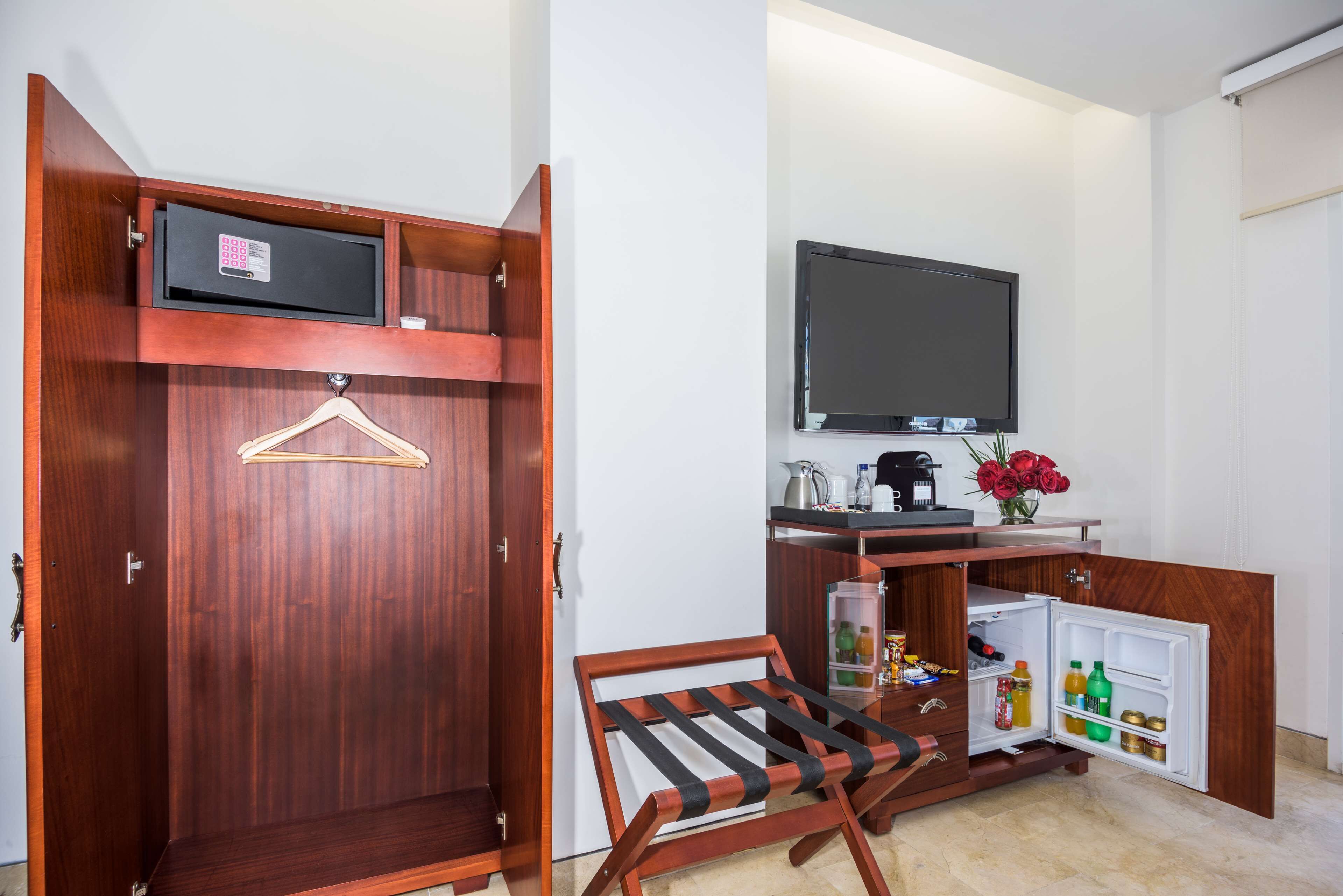 A compact room with a wooden wardrobe, a small table with a chair, a flat-screen TV, and a mini-fridge.