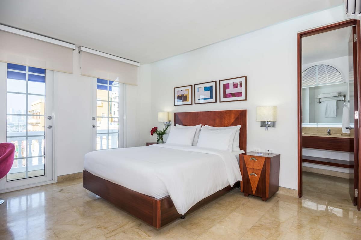 A hotel room with a large bed, a wooden headboard, a nightstand, a mirror, and a bathroom visible through an open door.