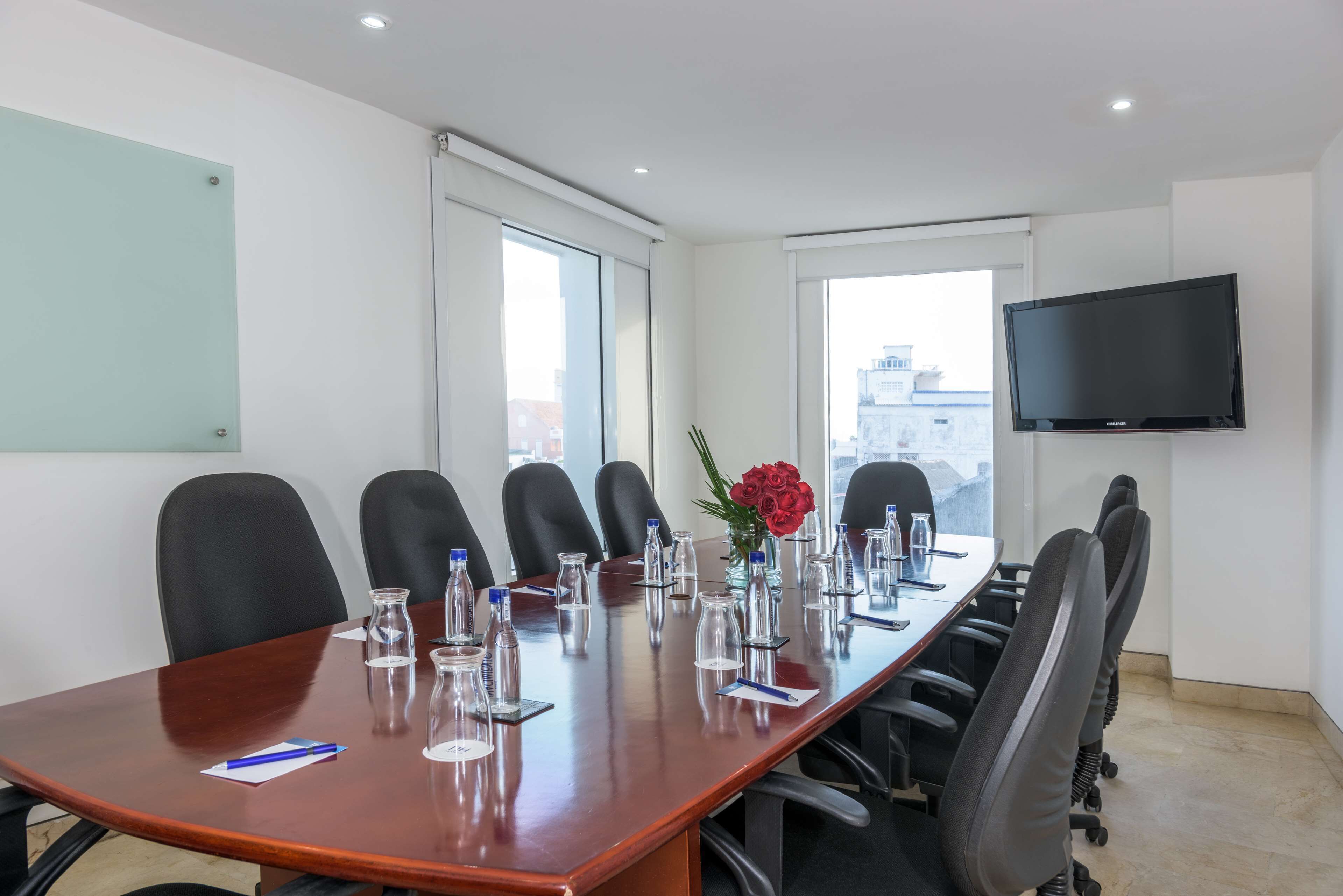A conference room with a long table, black chairs, a TV mounted on the wall, and large windows.