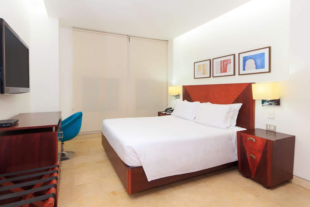 A hotel room with a bed, bedside tables, a desk, a chair, a TV, and artwork on the wall.