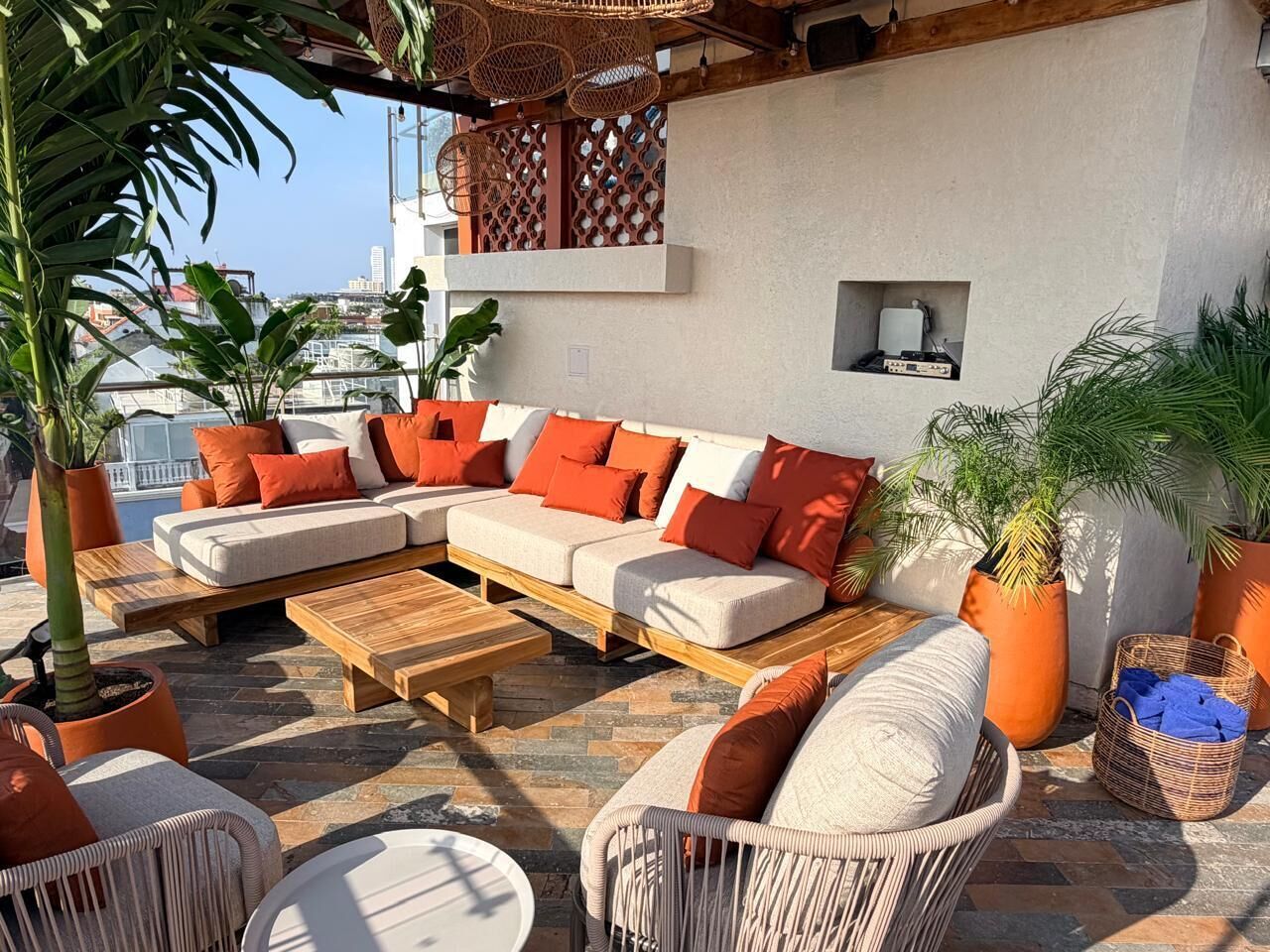 A rooftop lounge with a sectional sofa, a wooden coffee table, wicker chairs, and potted plants.