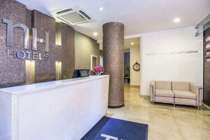Zona de recepción de hotel con un mostrador de mármol, un pilar grande, un sofá beige y un reloj en la pared.