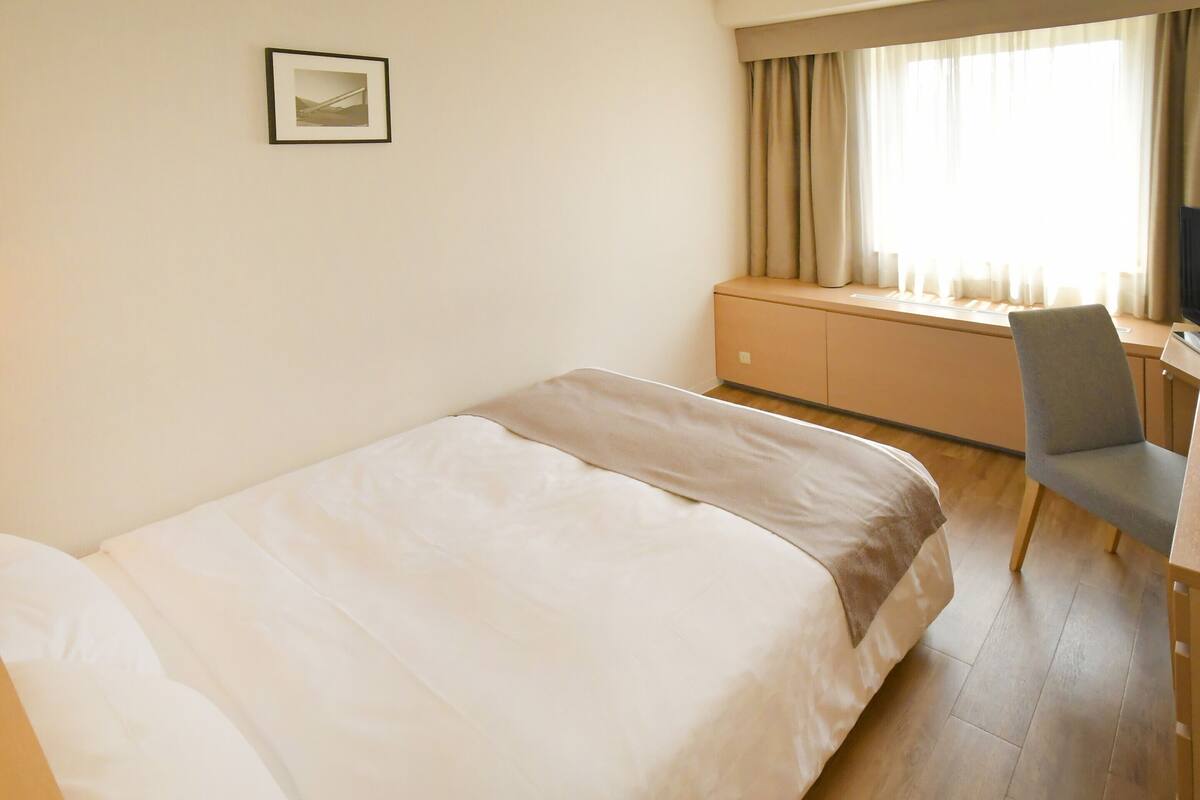 A hotel room with a bed, a desk, a chair, a window with curtains, and a framed picture on the wall.