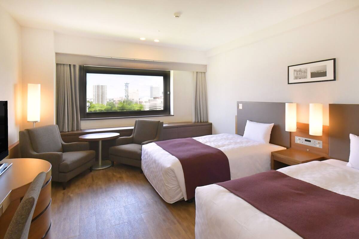 A hotel room with two beds, a desk, a chair, and a window with a city view.