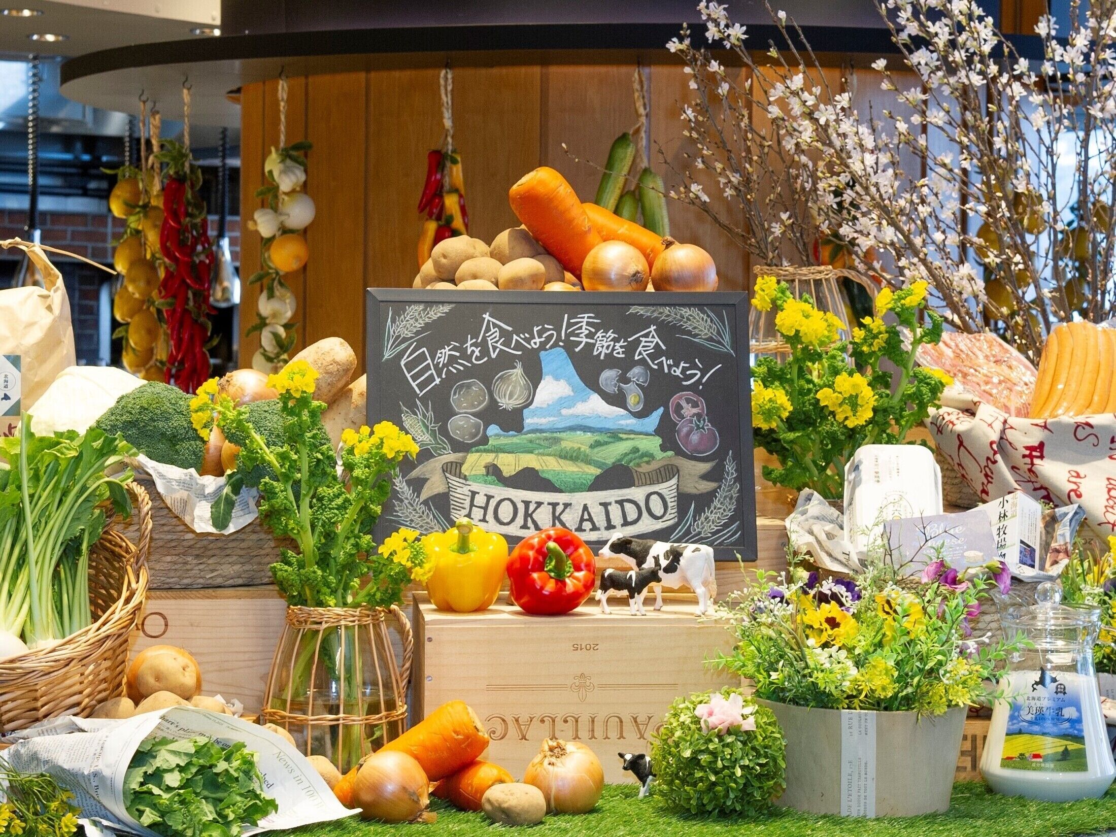 A display of fresh vegetables and fruits, including carrots, potatoes, onions, and bell peppers, with a chalkboard sign that reads "HOKKAIDO".