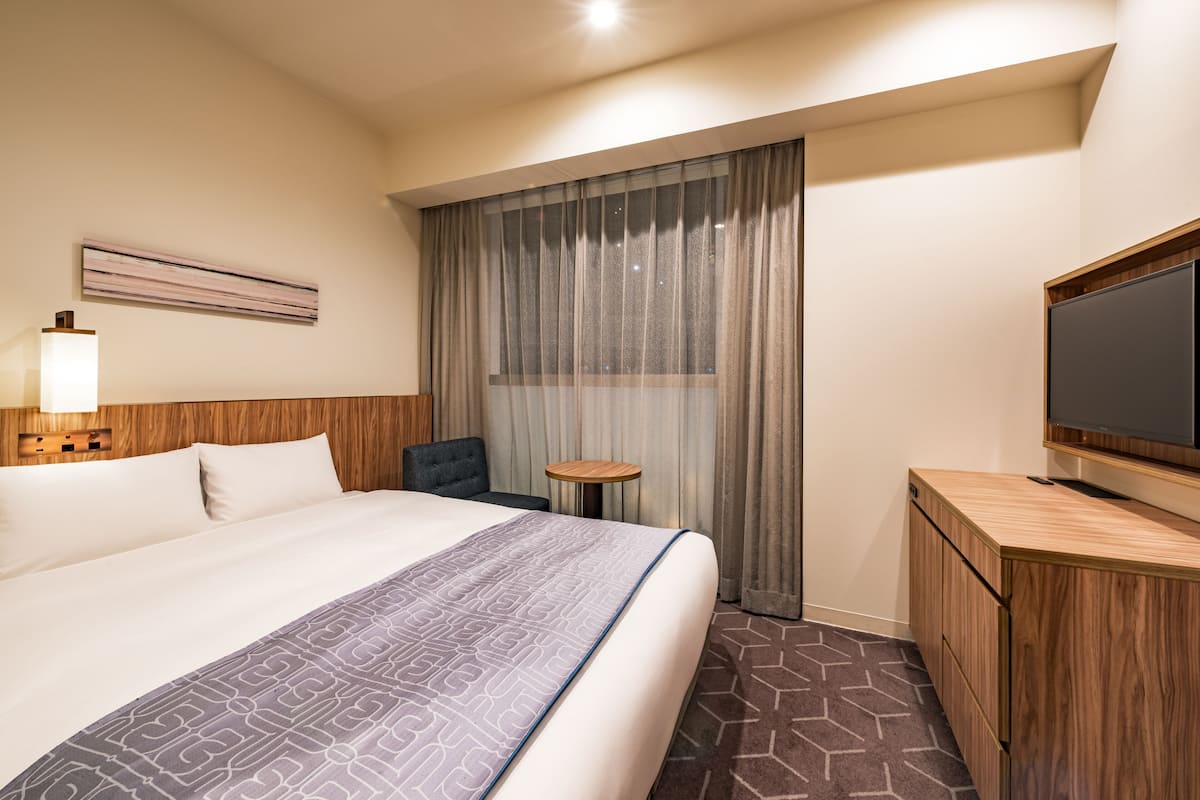 A hotel room with a large bed, a TV, a desk, and a window with curtains.