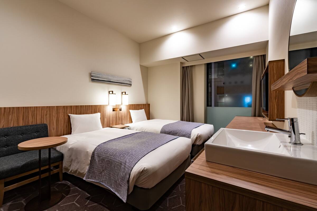 A hotel room with two beds, a sofa, a small round table, a large mirror, and a bathroom with a sink and mirror.