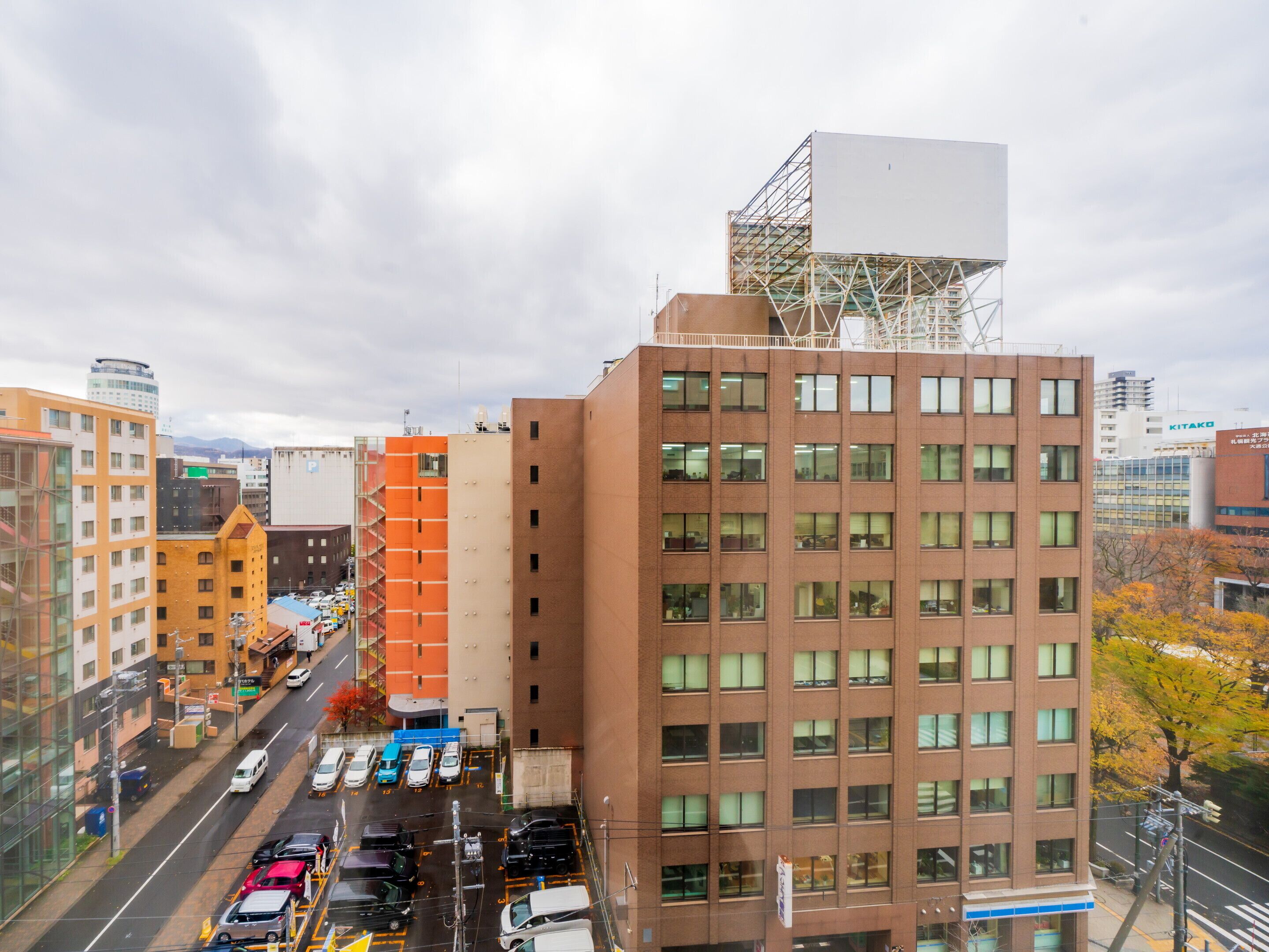 A multi-story building with a large billboard on top, surrounded by other buildings and a busy street with parked cars.