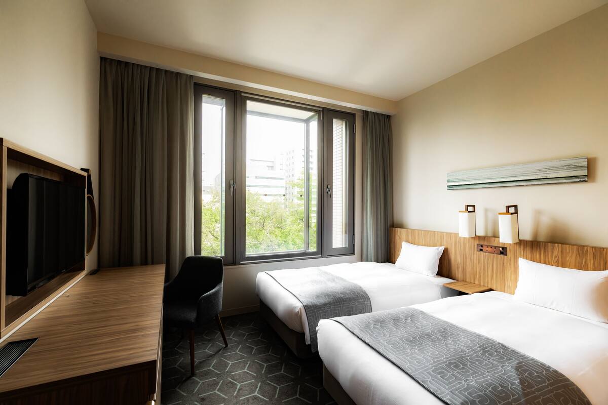 A hotel room with two beds, a desk, a TV, and a large window with curtains.