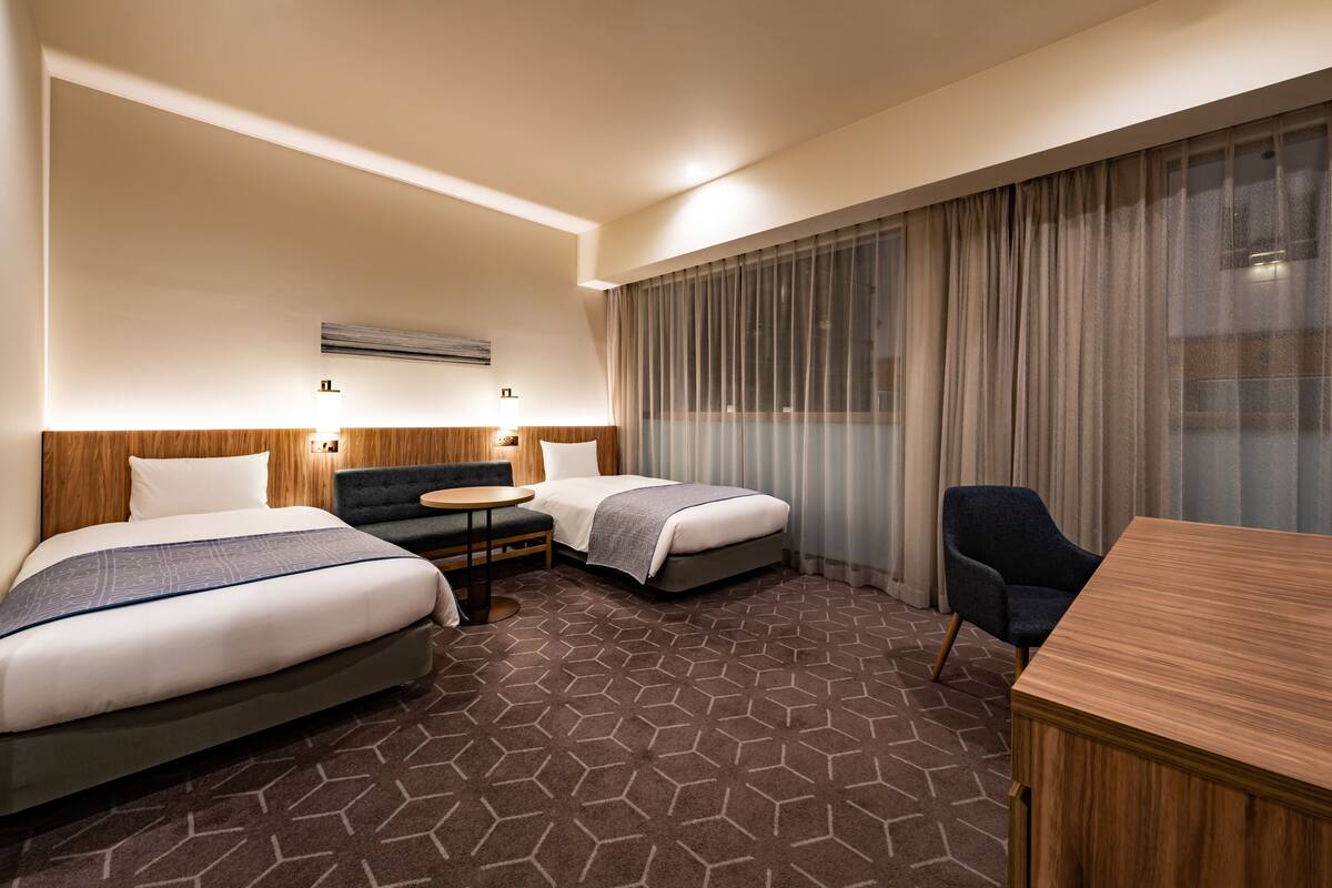 A hotel room with two beds, a desk, a chair, and a large window with curtains.