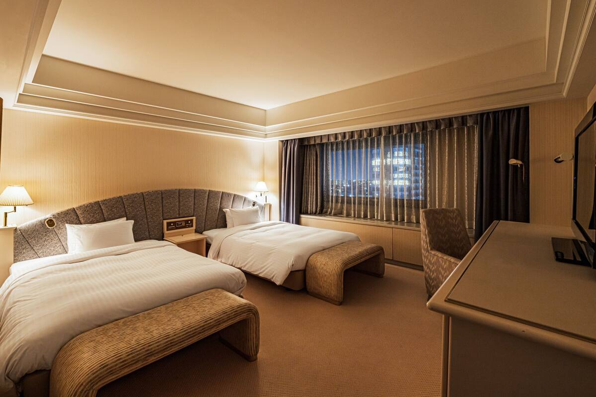 A hotel room with two beds, a desk, a chair, and a TV.