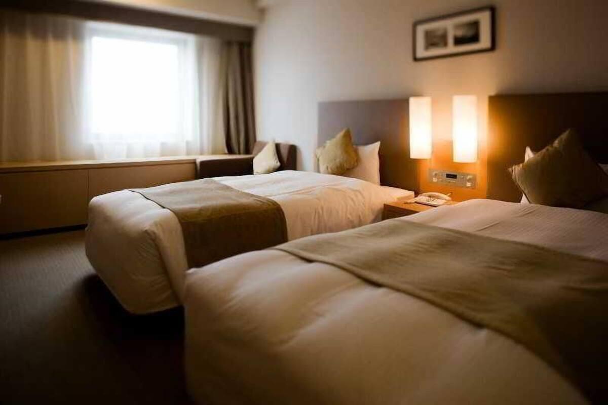 A hotel room with two beds, a bedside table with a lamp, a window with curtains, and a wall-mounted picture.