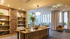 A modern hotel lobby with a central wooden reception desk, shelves with books and decorative items, a large window with a view of trees, and a cozy seating area with a sofa and armchairs.