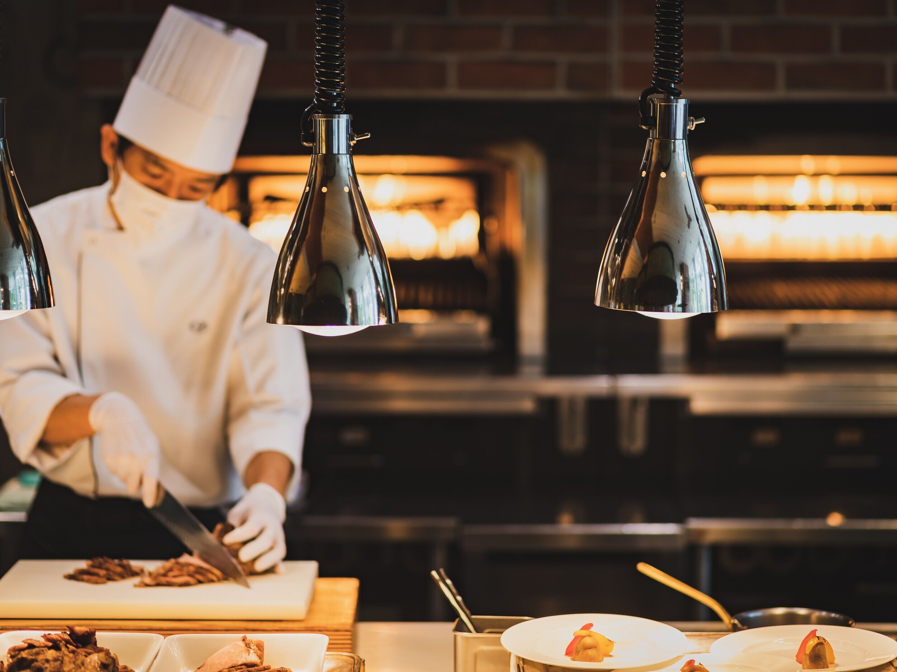 Un chef preparando comida en una cocina con lámparas colgantes y una pared de ladrillo al fondo.