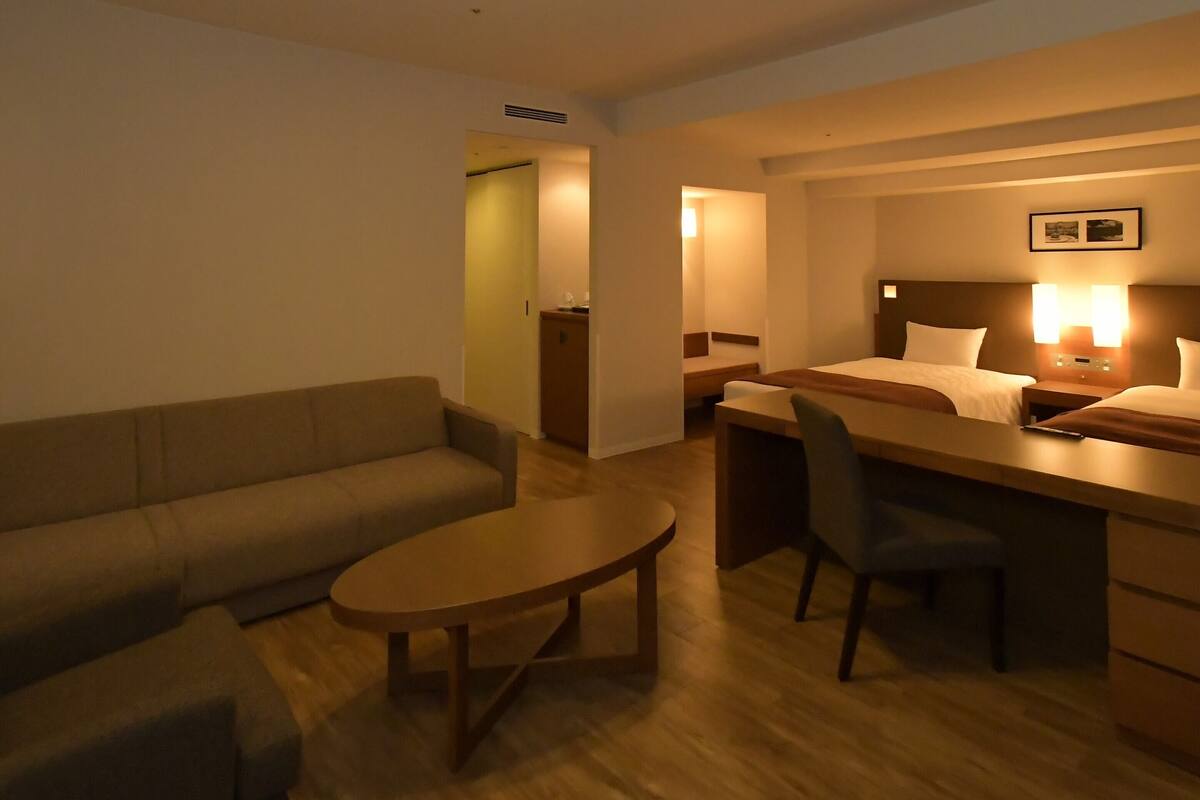A hotel room with a sofa, a small round table, a desk with a chair, a bed, and a nightstand with a lamp.