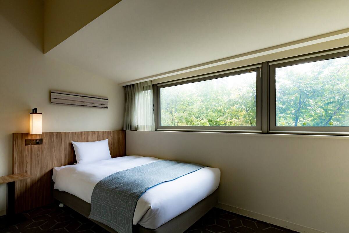 A hotel room with a bed, a nightstand, a wall-mounted lamp, and a window with a view of trees.