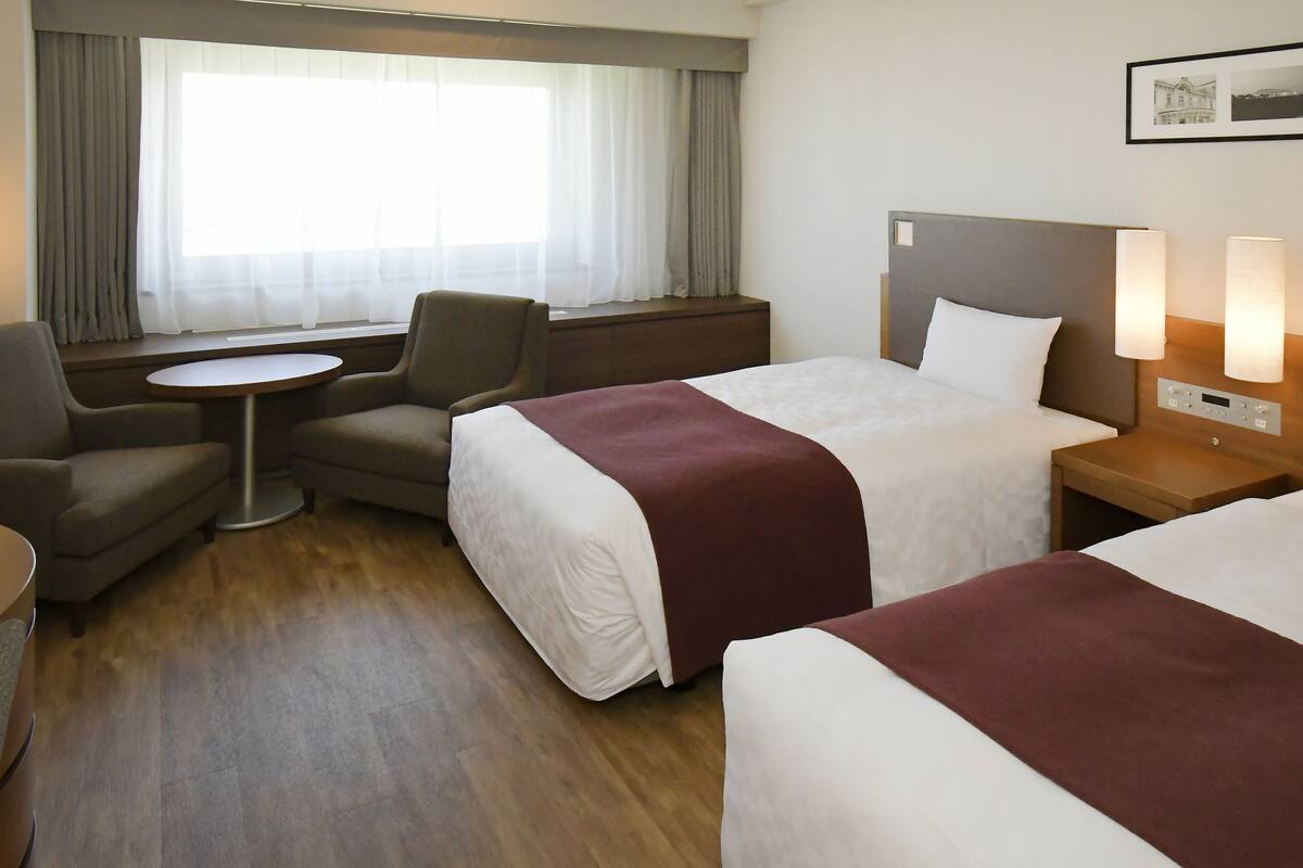 A hotel room with two beds, a small table, a chair, and a window with curtains.