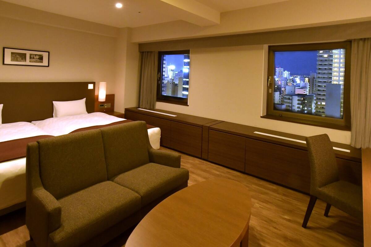 A hotel room with a bed, a sofa, a small table, and a view of a cityscape through two windows.
